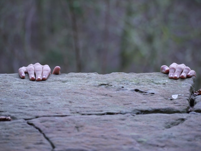 Two hands gripping the edge of a weathered stone ledge, set against a blurred forest background, suggesting clinging on to the ledge for dear life. Photo by Fiona Dodd on Unsplash
