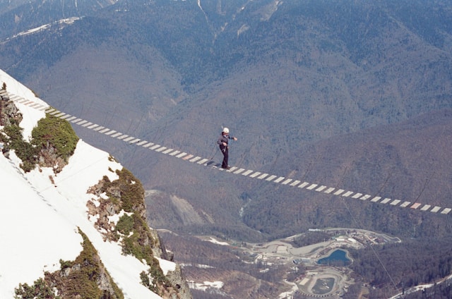 A person carefully walks on a narrow, high-altitude rope bridge over a mountainous landscape. Snowy slopes and a valley are visible below, conveying a sense of danger. Photo by Kamil Foatov on Unsplash
