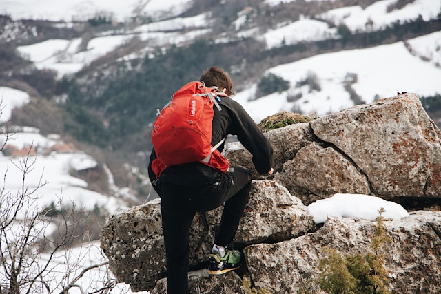 Young male with a red backpack climbing a snowy rock in a mountainous landscape. Photo by Mael Balland on Unsplash