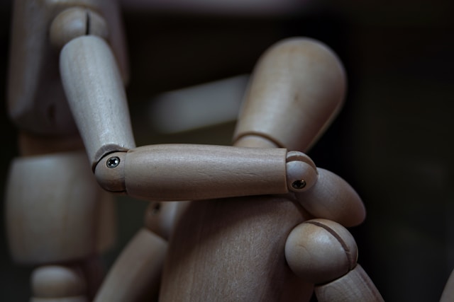 A wooden artist's mannequin standing next to a second seated wooden artist's mannequin. The first mannequin has its hand placed supportively on the shoulder of the second mannequin as if to offer comfort. The lighting is sombre, suggesting that the second mannequin is sad. Photo by Marco Bianchetti on Unsplash.